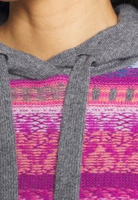 Huvtröja i grått tyg, med ett mönstrat kropp i nyanser av rosa och lila. Texturerad stickning och kontrasterande dragsnören.