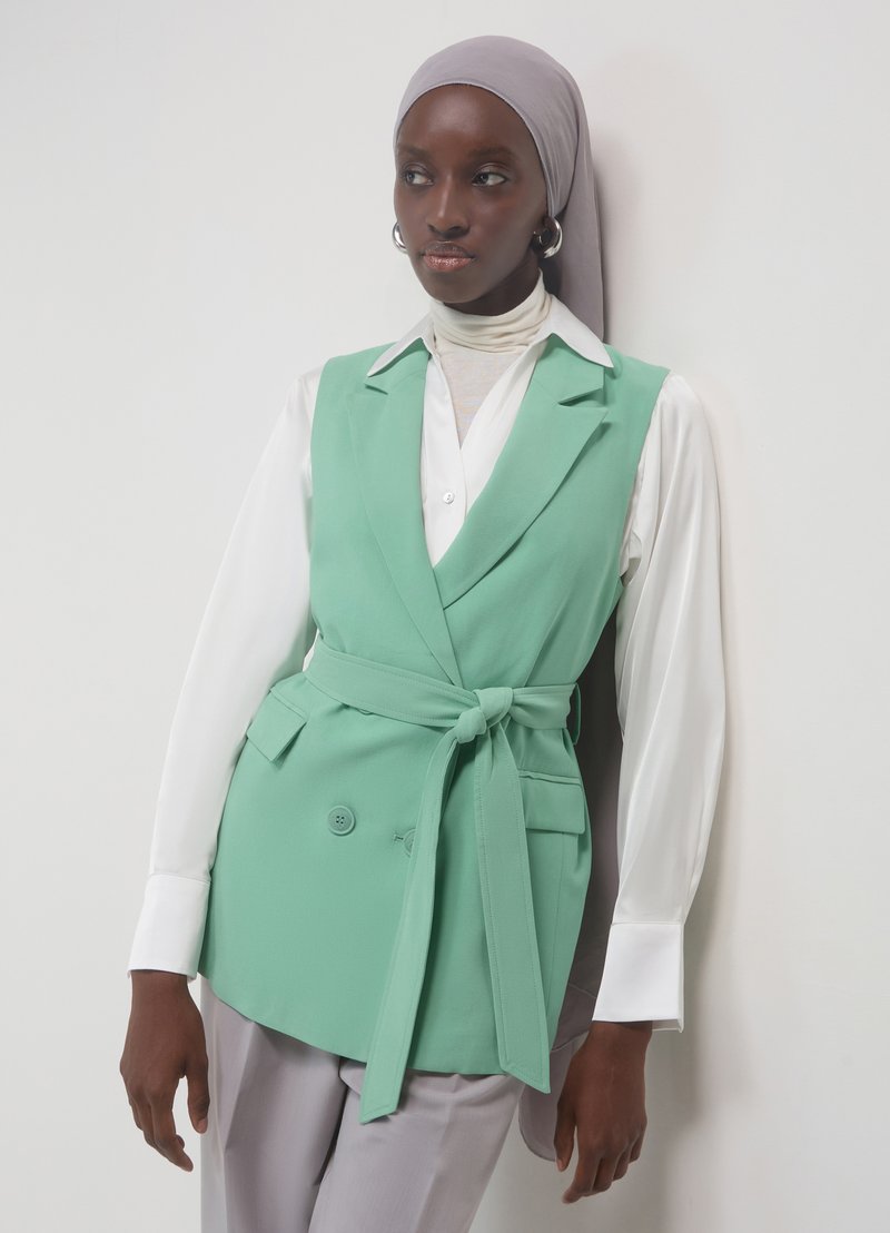 Minzgrüne Wickelweste mit doppeltem Knopfdesign, gebundener Taille und Taschen, kombiniert mit einer weißen Bluse. Das Model trägt ein hellgraues Kopftuch.