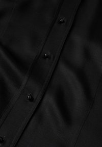 Camisa de tela negra con textura suave, que presenta una fila de botones negros redondos en la parte delantera y detalles de costura sutiles.