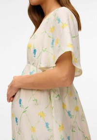 Robe de maternité florale avec de courtes manches volantées, ornée de fleurs jaunes et bleues sur un tissu beige clair, texture douce et coupe ample.