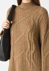 Ruskea neulepaita korkealla kauluksella, jossa on kuvioraitoja ja ribbitekstuuri. Musta olkalaukku nojaa paidan kylkeen.