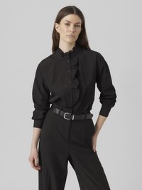 Camicia nera a maniche lunghe con colletto arricciato e pattina frontale, abbinata a pantaloni neri a vita alta e una cintura nera stretta con fibbia argentata.