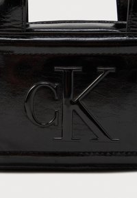 Czarna torebka z lakierowanej skóry z wypukłym logo "ck". Gładka tekstura z ostrymi krawędziami i usztywnionym designem, odpowiednia do codziennego użytku.