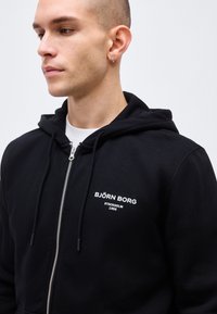 Junger Mann mit kurzen Haaren, der einen schwarzen Kapuzenpullover mit Reißverschluss trägt, auf dessen Brust der Schriftzug "BJÖRN BORG STOCKHOLM 1956" steht, und der nach links schaut.