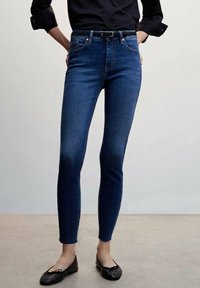 Woman wearing dark blue skinny jeans, black belt, black long-sleeve shirt with sleeves rolled, and black ballet flats, standing indoors.