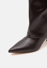 Botte haute en cuir lisse marron foncé avec bout pointu et petit talon sur fond clair.