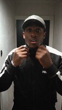 Fekete sportdzseki fehér csíkokkal, elülső cipzárral és logóval a mellkason. Sötétszürke sapka, a kezében a dzseki gallérját igazítja.