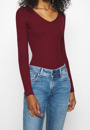 Femme portant un haut bordeaux côtelé ajusté à manches longues et un jean bleu taille haute sur fond uni.