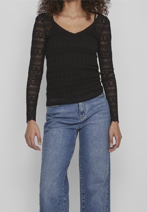 Kvinde iført sort blondebluse med lange ærmer og blå jeans, stående foran en ensfarvet lys grå baggrund.