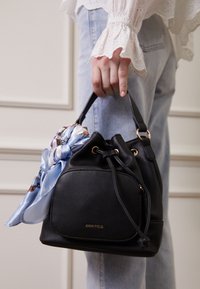 Schwarze Leder-Eimer-Tasche mit einem Zugverschluss, verziert mit einem hellblauen Seidenschal und goldfarbenen Beschlägen. Markenname an der Vorderseite sichtbar.