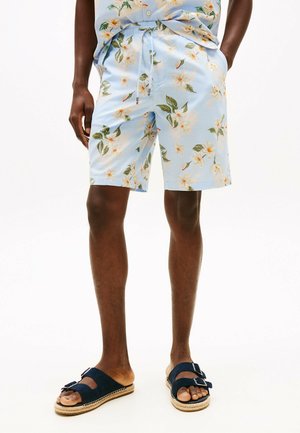 Shorts da uomo in blu chiaro con motivi floreali e foglie verdi. Caratterizzati da una vita elastica e un cordoncino. Abbinati a sandali blu navy con fibbie.