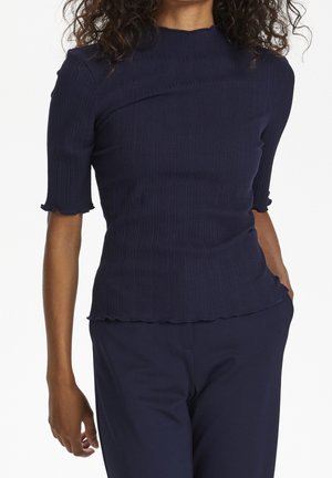 Top de punto acanalado azul marino con mangas cortas y bordes con ondas; diseño ajustado con cuello alto, textura suave y detalles mínimos.