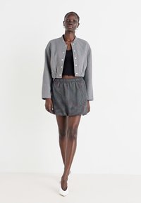 Veste grise courte avec des boutons-pression argentés, associée à un crop top noir et un short rayé à taille cintrée. Le modèle porte des talons blancs.