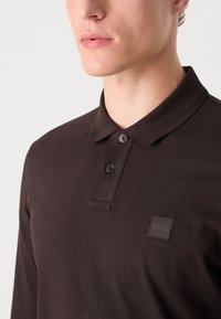 Brun langærmet polo shirt med en struktureret krave, to sorte knapper og et firkantet logo-patch på brystet. Glat stoftekstur.