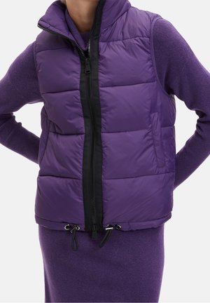 Osoba ubrana w fioletową pikowaną kamizelkę bez rękawów na fioletowej bluzce z długim rękawem i dopasowanej spódnicy, stojąca z rękami na biodrach.