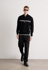 Μαύρο φλις πουλόβερ μισής ζώνης με λευκές οριζόντιες ρίγες, συνδυασμένο με μαύρο παντελόνι και παπούτσια. Μοντέλο που φοράει γυαλιά ηλίου. Λειαστή υφή.