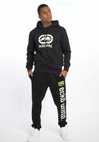 Sort hættetrøje med frontlogo og snor, parret med sorte sweatpants med et grønt logo og hvid tekst ned ad benet. Hvide sneakers.