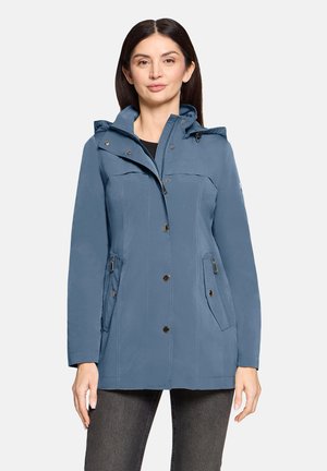 Kobieta w niebieskiej kurtce z kapturem, wyposażonej w guziki z przodu i zapinane na zamek boczne kieszenie, ubrana w ciemnoszare spodnie, stojąca na jednolitym tle.