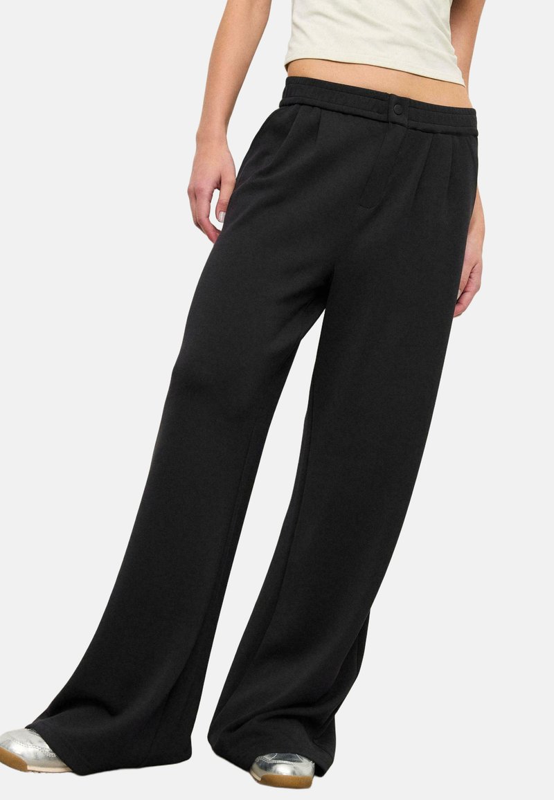 Personne portant un pantalon noir ample avec une ceinture plissée et des chaussures argentées, debout les mains détendues le long du corps.