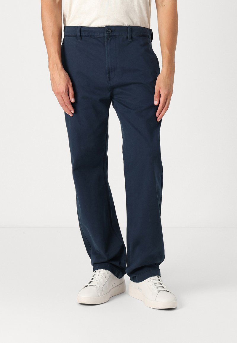 Chino marine avec une coupe droite, fabriqué en tissu mélangé de coton, doté d'une fermeture à bouton et de poches latérales, associé à des baskets blanches.