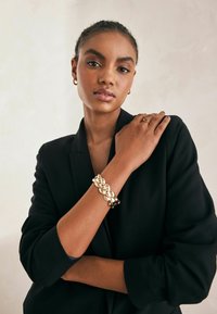 Bracelet en or tressé avec des éléments entrelacés. Porté au poignet, complété par un blazer noir. Texture lisse, finition brillante et design audacieux.