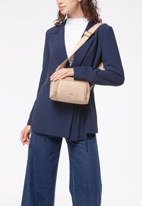Borsa a tracolla beige con superficie testurizzata, due scomparti con zip e una tracolla regolabile; indossata sopra un blazer blu scuro e jeans.