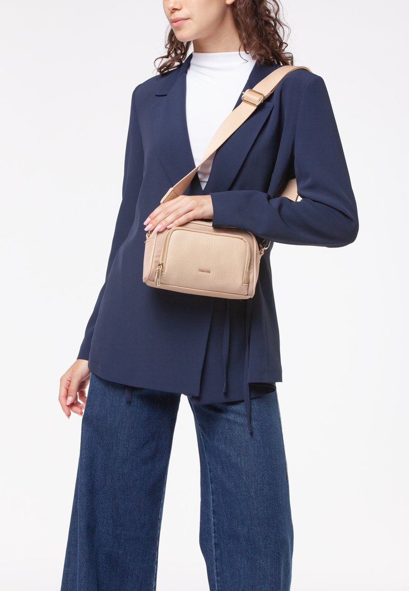 Borsa a tracolla beige con superficie testurizzata, due scomparti con zip e una tracolla regolabile; indossata sopra un blazer blu scuro e jeans.