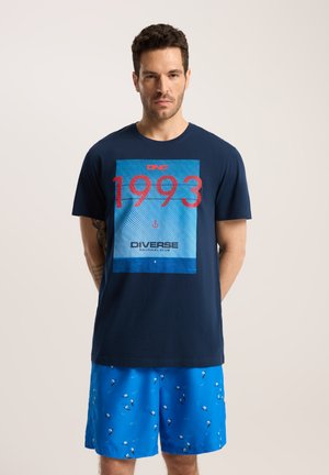 Granatowa koszulka z bawełny z wyraźnym czerwonym i niebieskim wzorem z napisem "1993" i "D'VERSE". Połączona z jasnoniebieskimi szortami z wzorem.