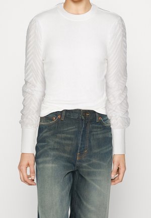 Personne portant un haut blanc à manches longues avec des manches transparentes texturées, associé à un jean bleu délavé taille haute, sur un fond uni.