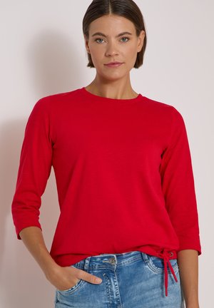Femme aux cheveux courts et foncés portant un haut rouge à manches longues avec un nœud sur le côté et un jean bleu, debout devant un fond clair.