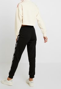 Zkrácená krémová mikina s červenými akcenty spárovaná s černými tepláky s bočním logem; modelka obutá v bílých teniskách, pohled zezadu.