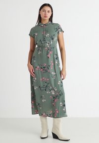 Robe maxi florale verte à manches courtes, en tissu doux, taille élastique et col boutonné. Portée avec des bottes à talons blanches.