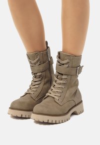 Botas de tobillo de ante verde oliva con cordones en la parte delantera, cremallera lateral y una correa con hebilla. Suela de goma gruesa con textura para un buen agarre.