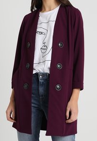 Burgundy dubbelknäppt kappa i mjukt tyg, med stora svarta knappar, sidofickor och uppvikta ärmar, bärs över en vit grafisk t-shirt.