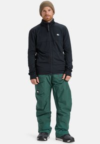 Giacca in pile nera con zip e colletto alto, abbinata a pantaloni larghi impermeabili verdi. Presenta toppe con logo e dettagli in gomma.