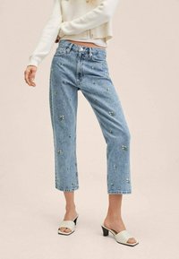 Jeans en denim bleu clair avec une coupe décontractée, ornés d'applications florales et d'une longueur courte. Assortis à des sandales à talons ouverts de couleur crème.