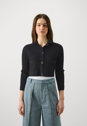 Cardigan cropped a coste nera con chiusura a bottoni, tasche frontali e colletto decorativo. Abbinato a pantaloni plissettati di colore azzurro chiaro.