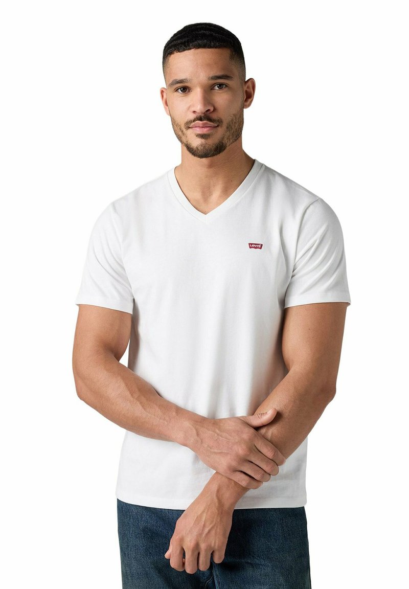 Mann mit kurzen Haaren und Bart, der ein weißes Levi's V-Ausschnitt T-Shirt und blaue Jeans trägt, steht mit verschränkten Armen über den Handgelenken.