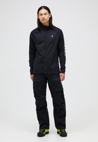 Mørkeblå mandlig zip-up jakke med lange ærmer, der har et logo og tekstdetalje. Sammen med sorte cargo bukser og sko med gule accenter.