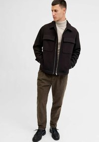 Svart zip-up jacka med två bröstfickor, kombinerad med bruna texturerade byxor och svarta snörskor. Modellen står mot en neutral bakgrund.