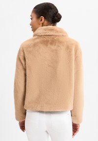 Beżowa kurtka z futra sztucznego z wysokim kołnierzem, luźnym krojem i miękką teksturą. Posiada prosty dół i długie rękawy.