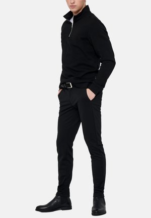 Mann trägt schwarzen Pullover mit halbem Reißverschluss, schlanke schwarze Hose mit Gürtel und schwarze Leder-Stiefeletten, Hände in den Taschen, vor weißem Hintergrund.