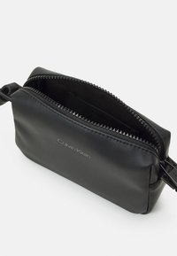 Bolsa de cuero negro con cremallera y textura suave, que presenta un logo plateado y un detalle de cremallera en contraste. El interior es oscuro y sin adornos.