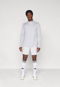 Αθλητικό ανοιχτό γκρι τοπ με φερμουάρ στο τέταρτο, συνδυασμένο με αντίστοιχα σορτς, λευκές κάλτσες με μαύρες ρίγες και λευκά ποδοσφαιρικά παπούτσια.