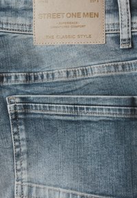 Poche arrière d'un jean en denim bleu clair avec un patch en cuir beige portant l'inscription "Street One Men" et passant de ceinture cousu.
