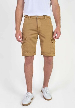 Homme portant un short cargo couleur camel retroussé, des baskets blanches et un t-shirt blanc, se tenant devant un fond clair.