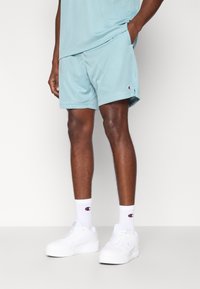 Vaaleansiniset verkkokankaasta valmistetut urheilushortsit, joissa on nyörityskaistale vyötäröllä, sivutaskut ja pieni logopistokke. Yhdistetty valkoisiin lenkkareihin ja sukkiin.