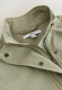 Col col en denim vert clair avec fermeture éclair partiellement ouverte et boutons-pression métalliques, montrant une étiquette de vêtement blanche à l'intérieur.