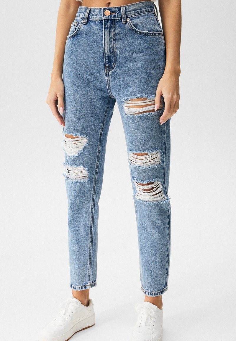 Jeans de cintura alta de color azul claro con desgastes rasgados, dobladillo deshilachado y un ajuste relajado. Combinados con zapatillas blancas.