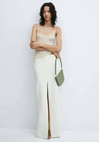 Bežový rebrovaný top na ramienkach spárovaný s bielou sukňou až na zem s bočným rozparkom. Modelka drží malú zelenú kabelku so štruktúrovaným dizajnom.